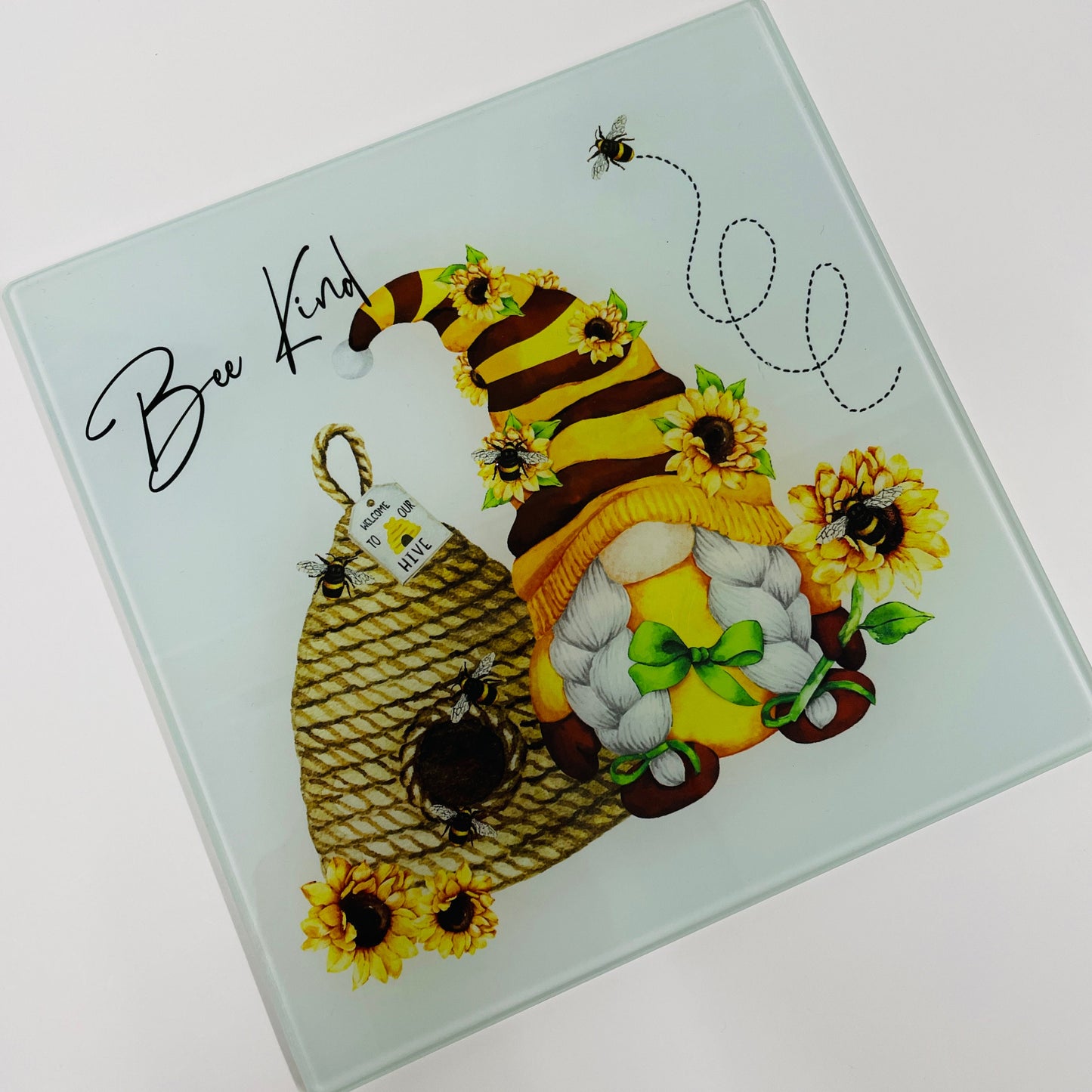 This is a 20cm square glass chopping board. Printed on the reverse of the board is a bee hive with bees flying around and a bee themed gonk with plaits. the colour theme is yellow, orange and brown. There are sunflowers on the gonks hat.