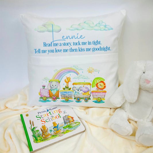 This is a white cushion cover with a pocket on the front. The pocket is to store a book. At the top is a name and the phrase, read me a story, tuck me in tight, tell me you love me then kiss me goodnight. On the front of the pocket is a little train full of animals riding past a rainbow.