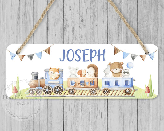 This is a white wooden nursery door sign with jute rope to hang anywhere. This design has a blue animal theme with a fox, mouse, hedgehog, squirrel, rabbit, bear and raccoon riding a train. At the top is bunting and a name. Nursery or bedroom decor