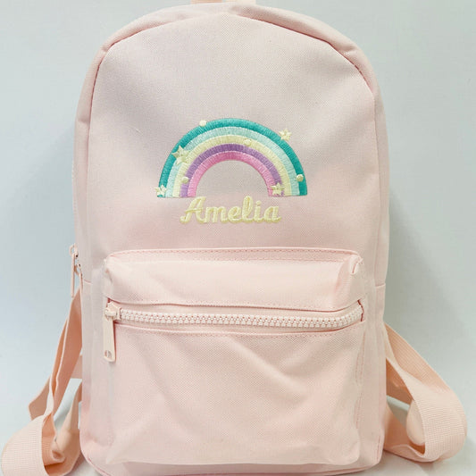 A light pink backpack with a pastel rainbow embroidered on the front. Below the rainbow is a name also embroidered. The bag has a main zip compartment and also a front pocket. With fully adjustable straps these are great for school or nursery.